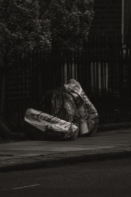 An overflowing collection of various waste items is displayed on a paved surface in front of a row of waste bins at a parking area in an urban setting. The heap includes cardboard boxes, plastic bags,