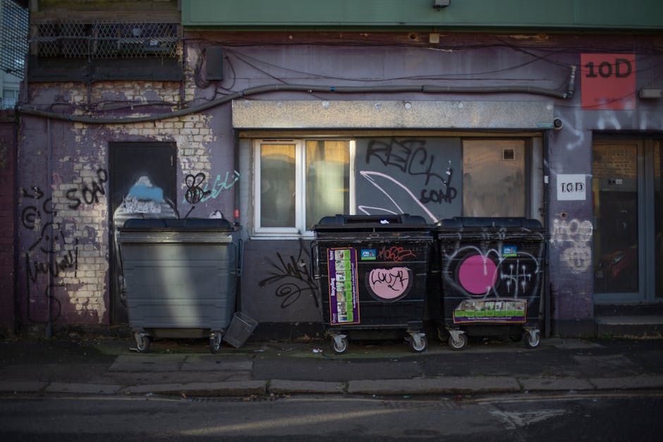 A photograph depicting the exterior of a weathered urban building with a purple brick façade, featuring a small white-framed window and graffiti tags in black, pink, and white spray paint. In front o